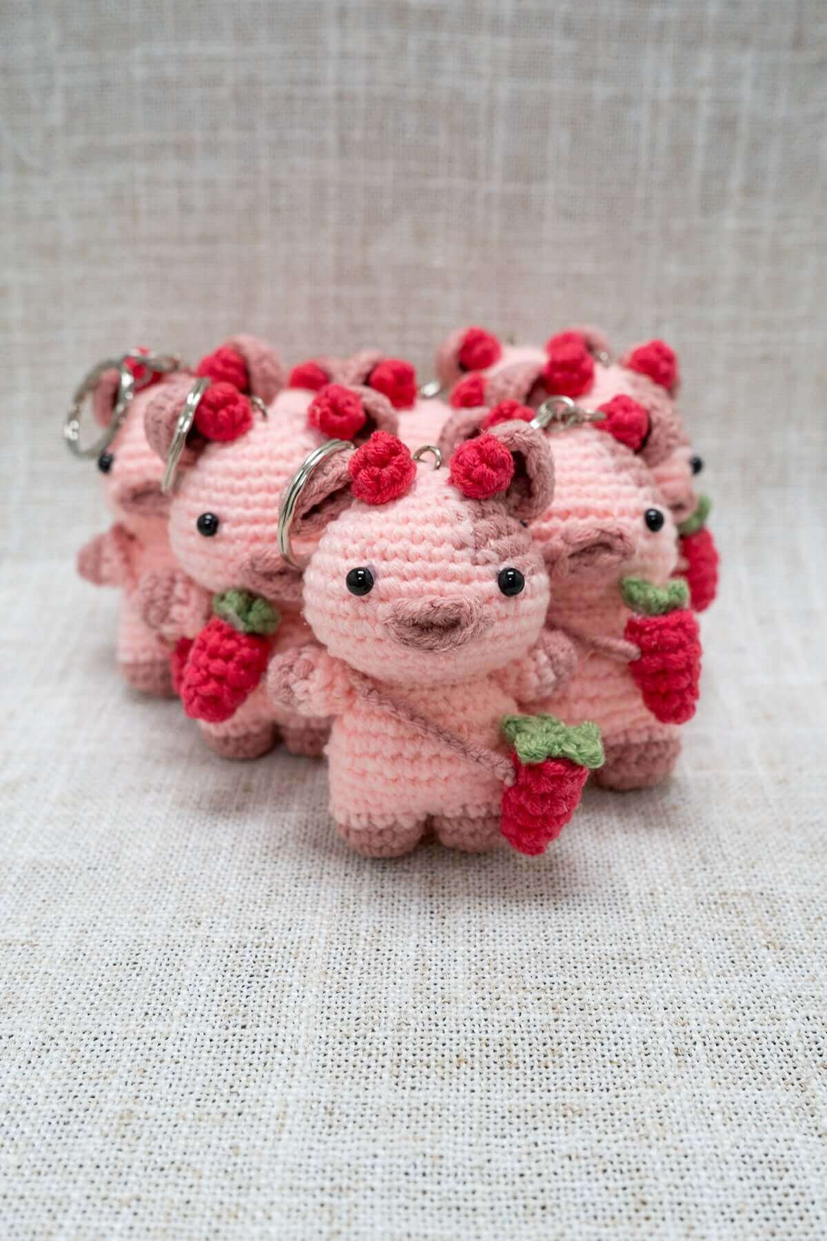 Group of handmade pink crochet bear keychains holding red berries on textured fabric background
