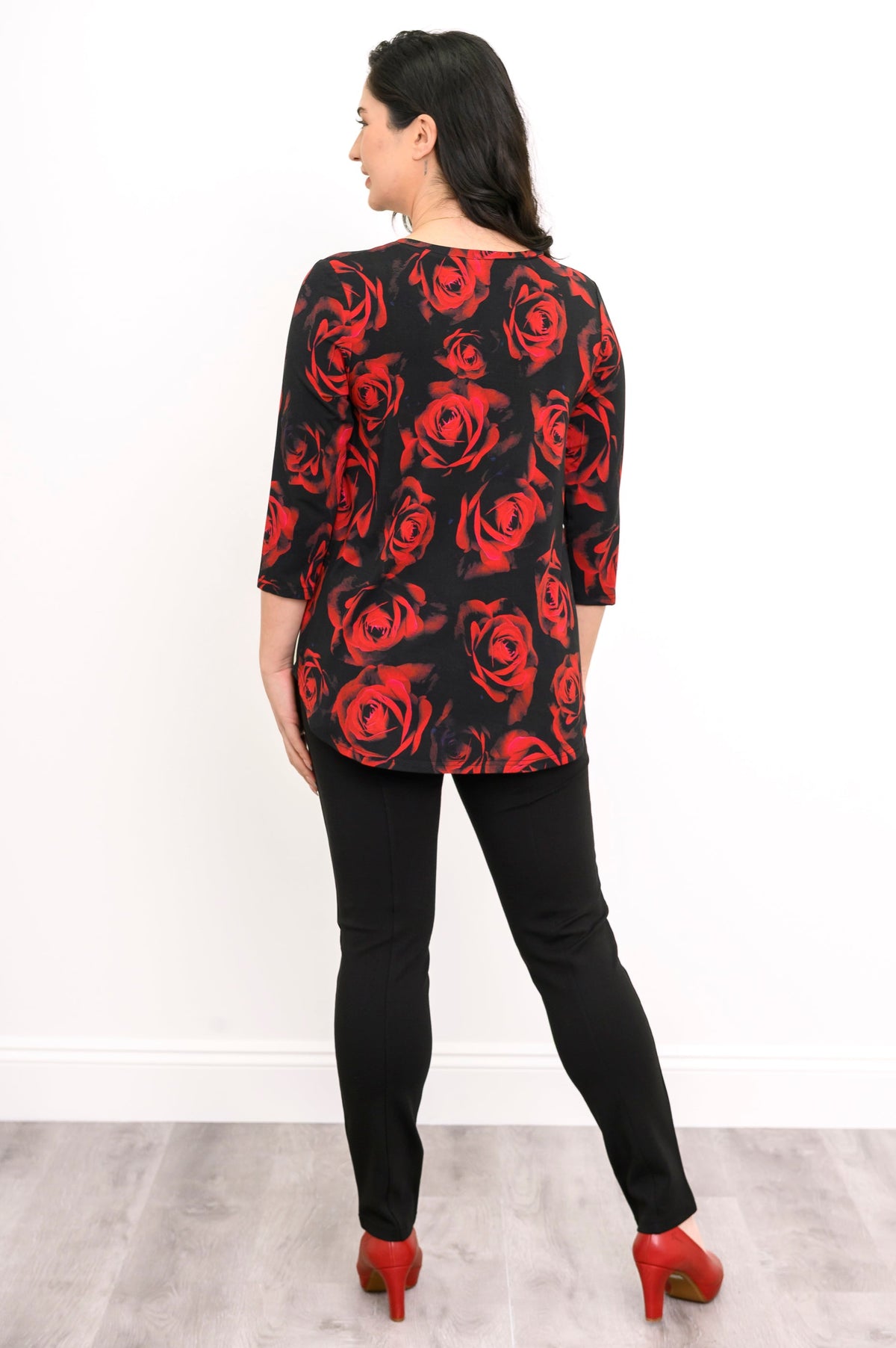 Woman wearing a black top with red floral pattern and black pants against a white background, back view