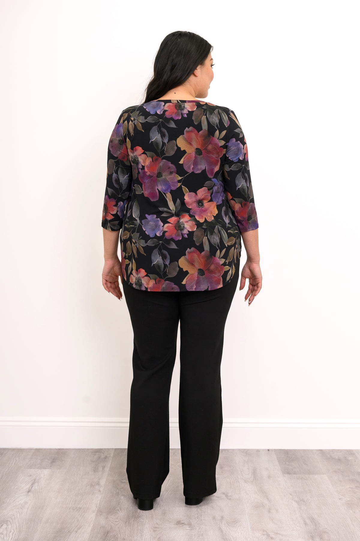 Woman wearing a multicolor floral patterned top and black pants on a white background, back view