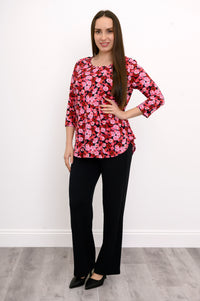 Model wearing a vibrant pink-red floral print A-line tunic paired with slim black trousers, standing front-facing.