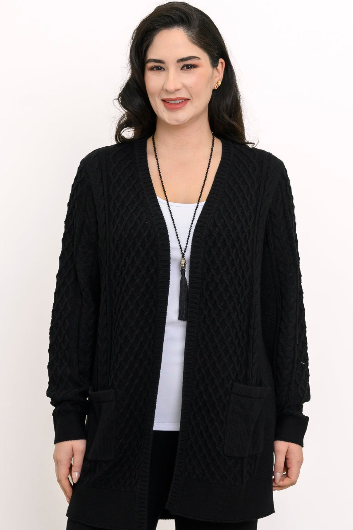 Close up front view of a woman wearing a black cable knit cardigan over a white top and black trousers.