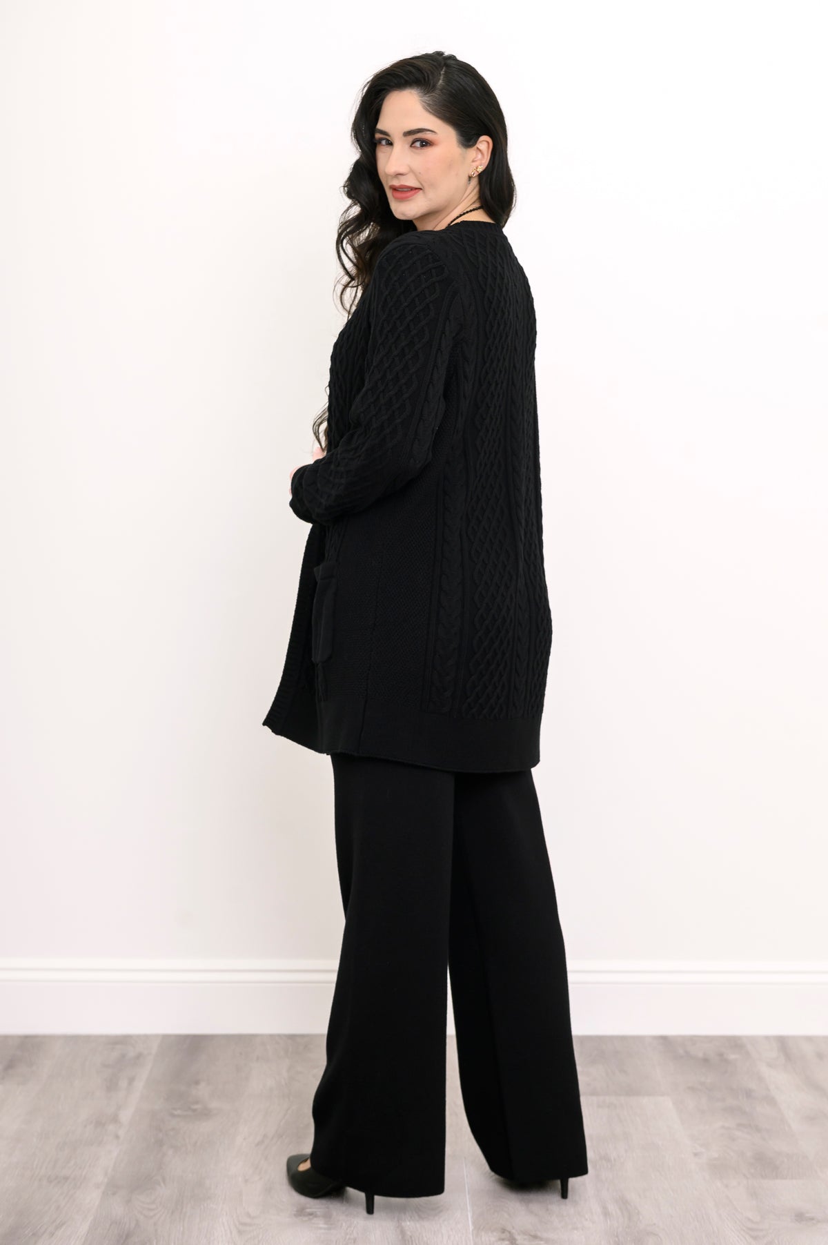 Side and rear view of a woman wearing a hip length black cable knit cardigan showing clean shoulder tailoring.