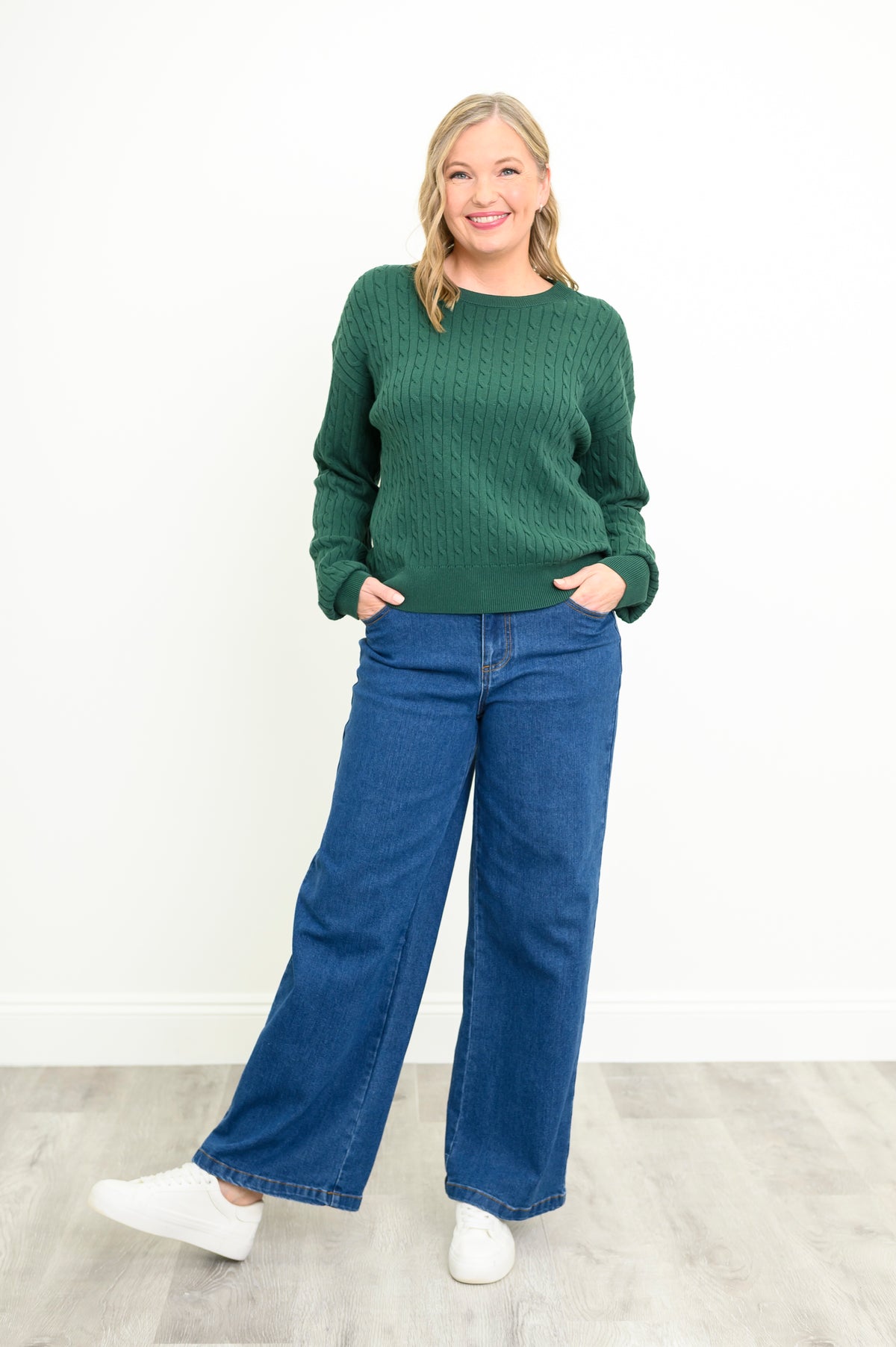 Model posing in a deep sage green cable-knit crewneck sweater and classic blue denim jeans, front-facing view.