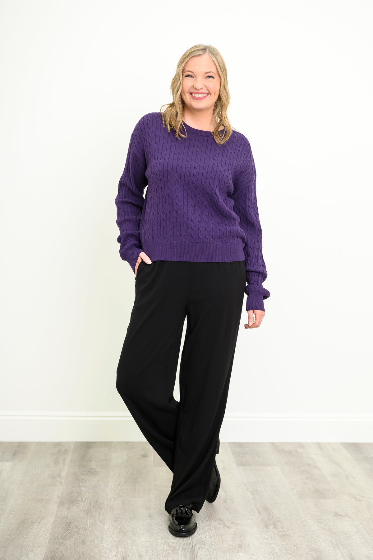 Smiling model in a cozy violet cable-knit pullover and black pants, posing naturally on a white background.
