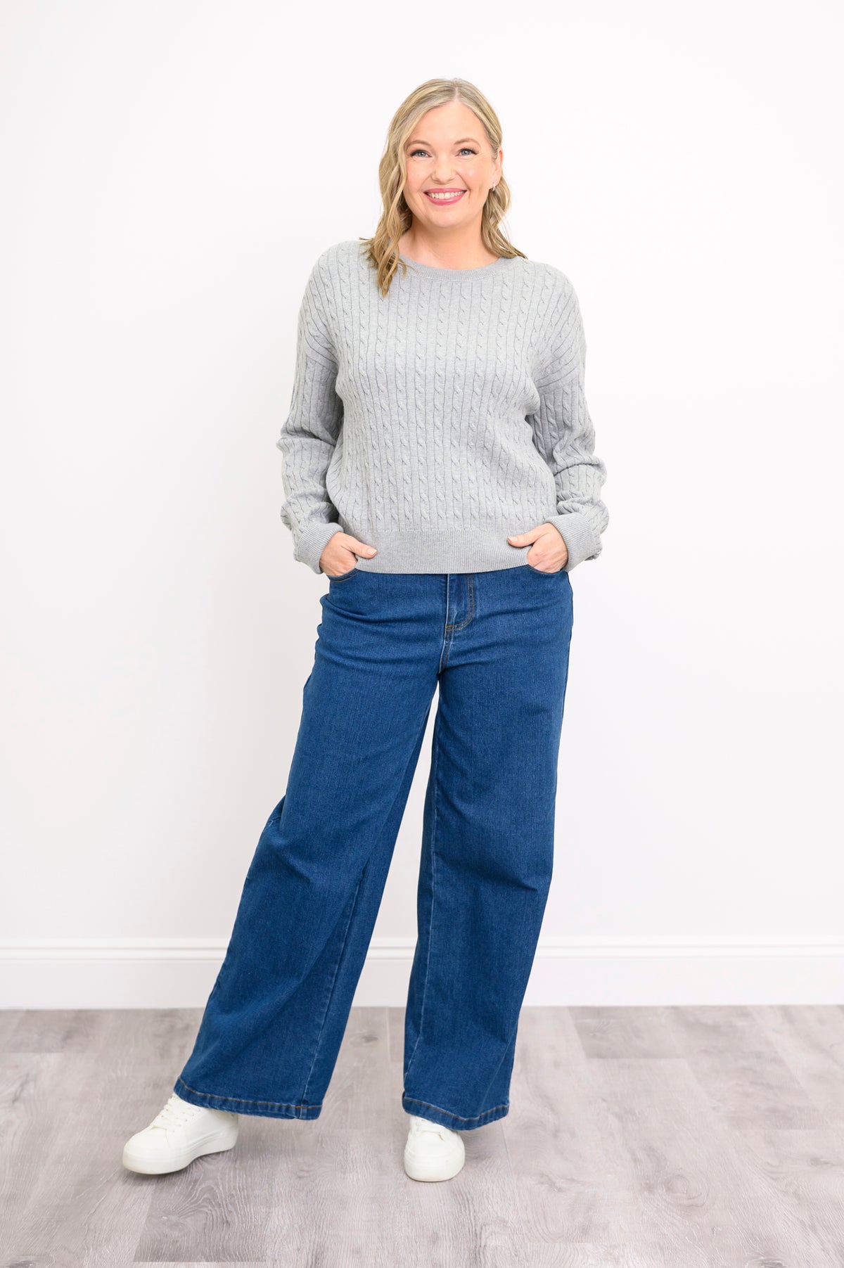 Smiling model in a cozy ash-colored cable-knit pullover and jeans, posing naturally on a white background.