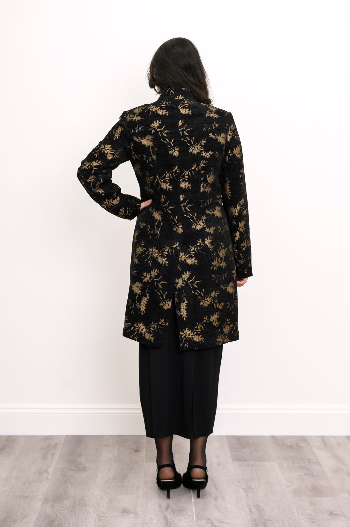 Back view of woman wearing long wool coat in black with gold leaf pattern and split hem