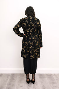 Back view of woman wearing long wool coat in black with gold leaf pattern and split hem