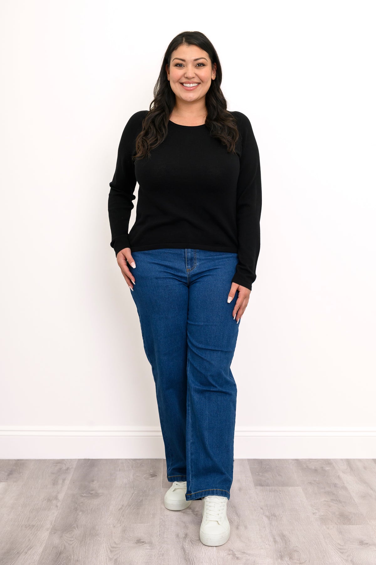 Model posing in an onyx scoop neck sweater featuring extended coverage sleeves, styled with casual blue denim pants.