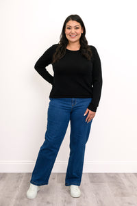 Front view of plus size female model wearing a black long-sleeve sweater featuring clean flat-finished wrist cuffs, hand on hip, with denim pants and white shoes. Against a white wall and gray floor