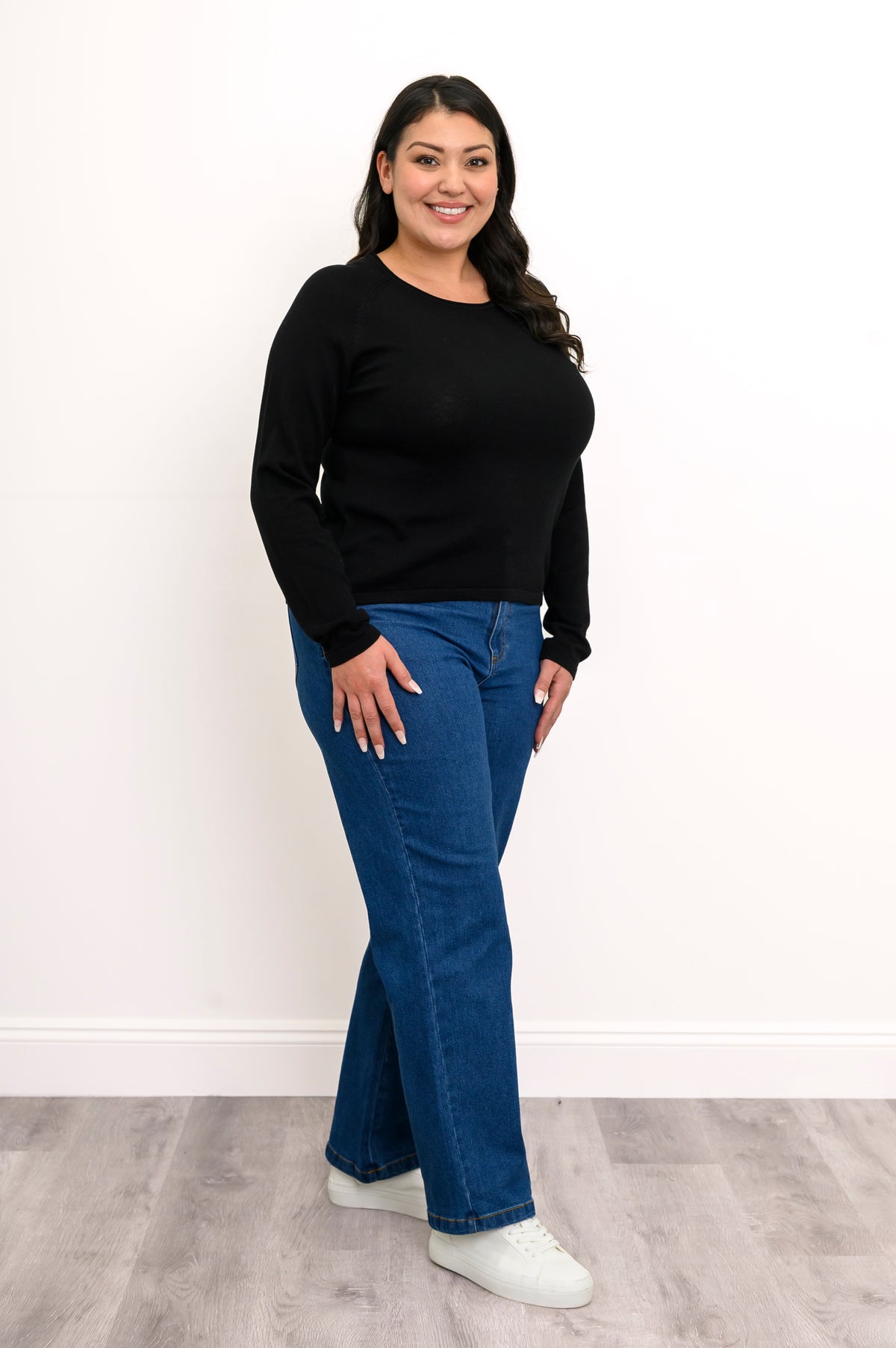 Side-facing view of a plus size female model wearing a black sweater with denim pants and white shoes, against a white wall and gray floor