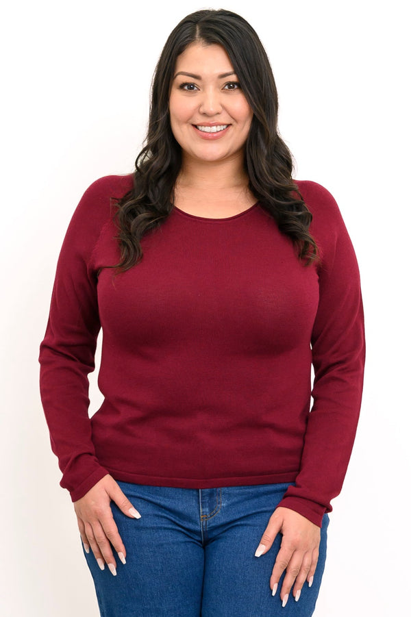 Front view of plus sized female model wearing a wine-red sweater with a scooped neckline and long sleeves with jeans. Styled against white backdrop