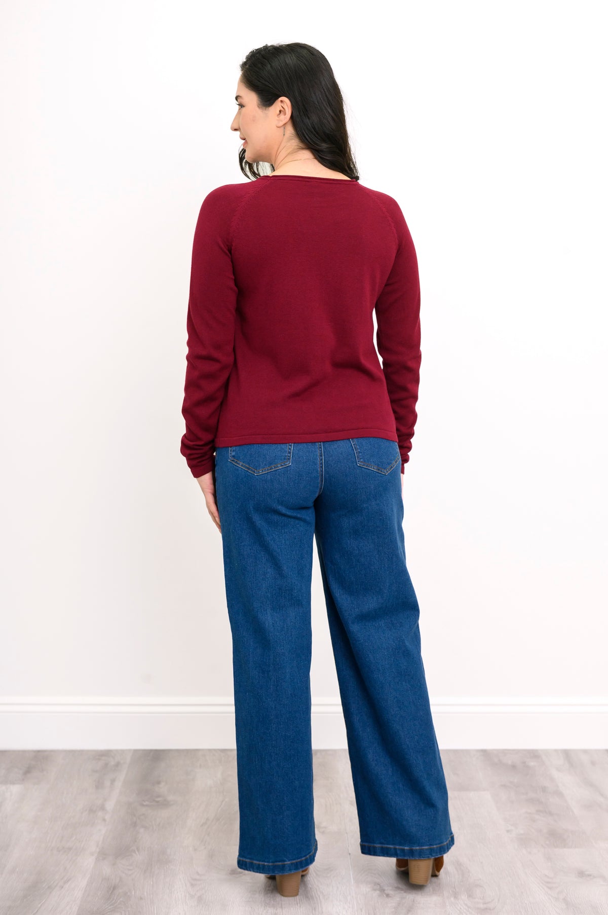 Back view of a women's long-sleeve dark red knit top, styled with blue jeans against a plain white backdrop.