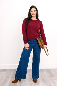Deep maroon knit pullover with long raglan sleeves and a rounded hem, shown on a model standing against a white wall.