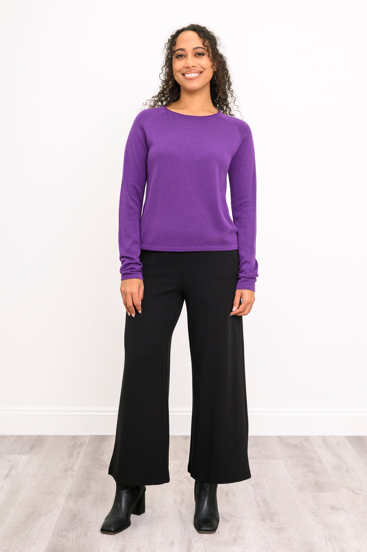 Aubergine knit pullover with long raglan sleeves and a rounded hem, shown on a model standing against a white wall.