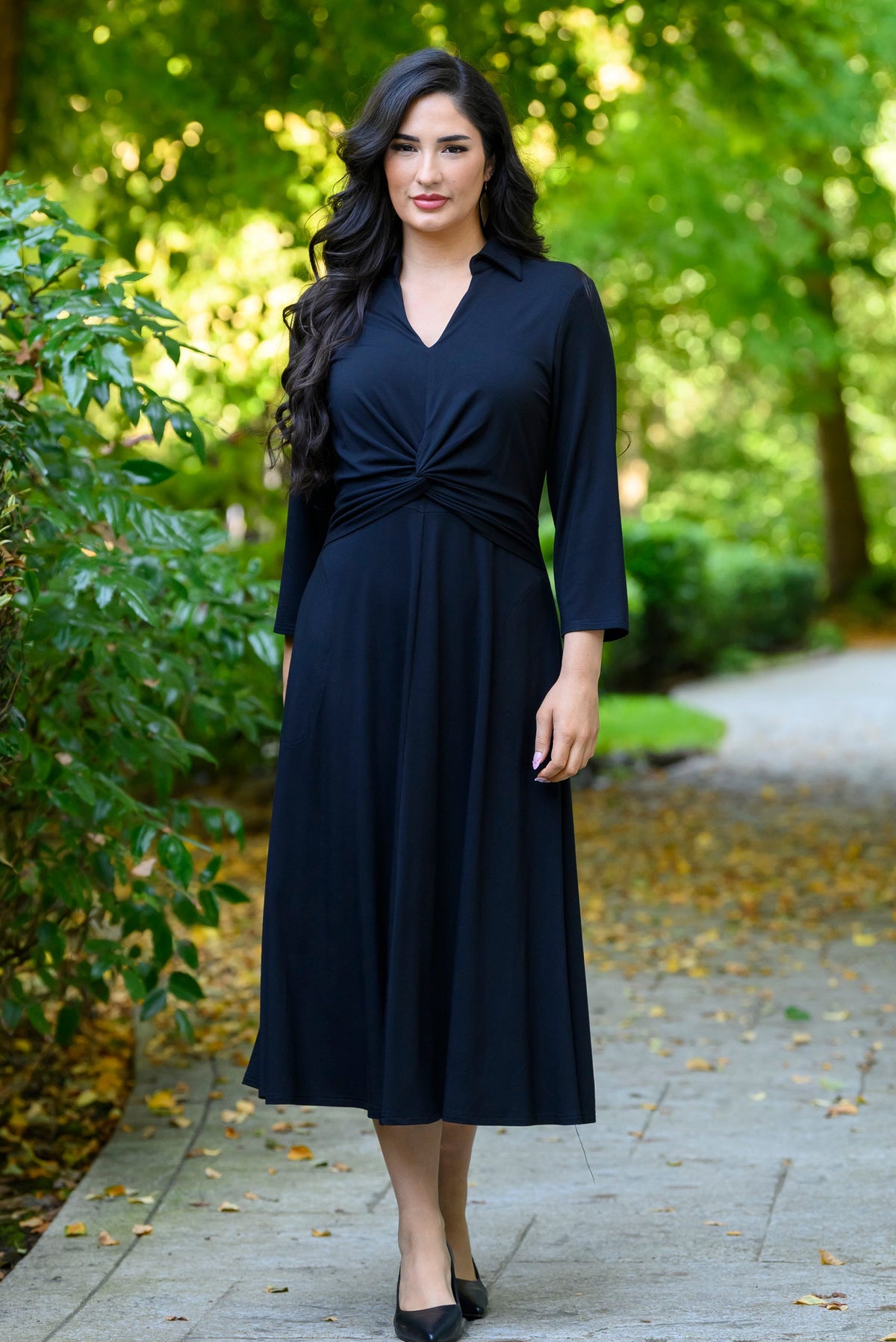front view of female model wearing a black midi wrap dress on a paved path in a lush green park.