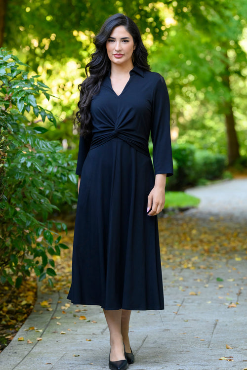 front view of female model wearing a black midi wrap dress on a paved path in a lush green park.