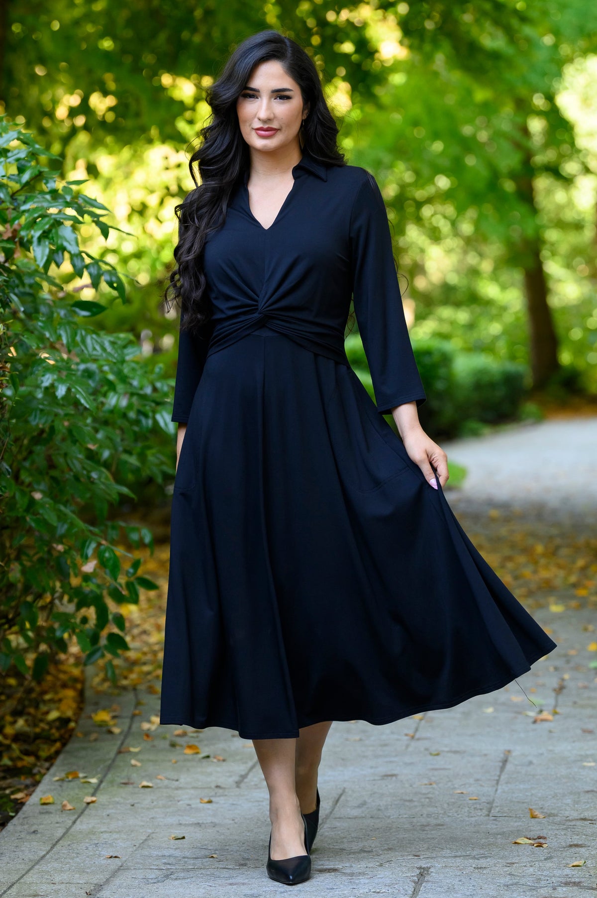front view of female model wearing a black midi dress with three-quarter sleeves, holding her dress on one side, in a green park