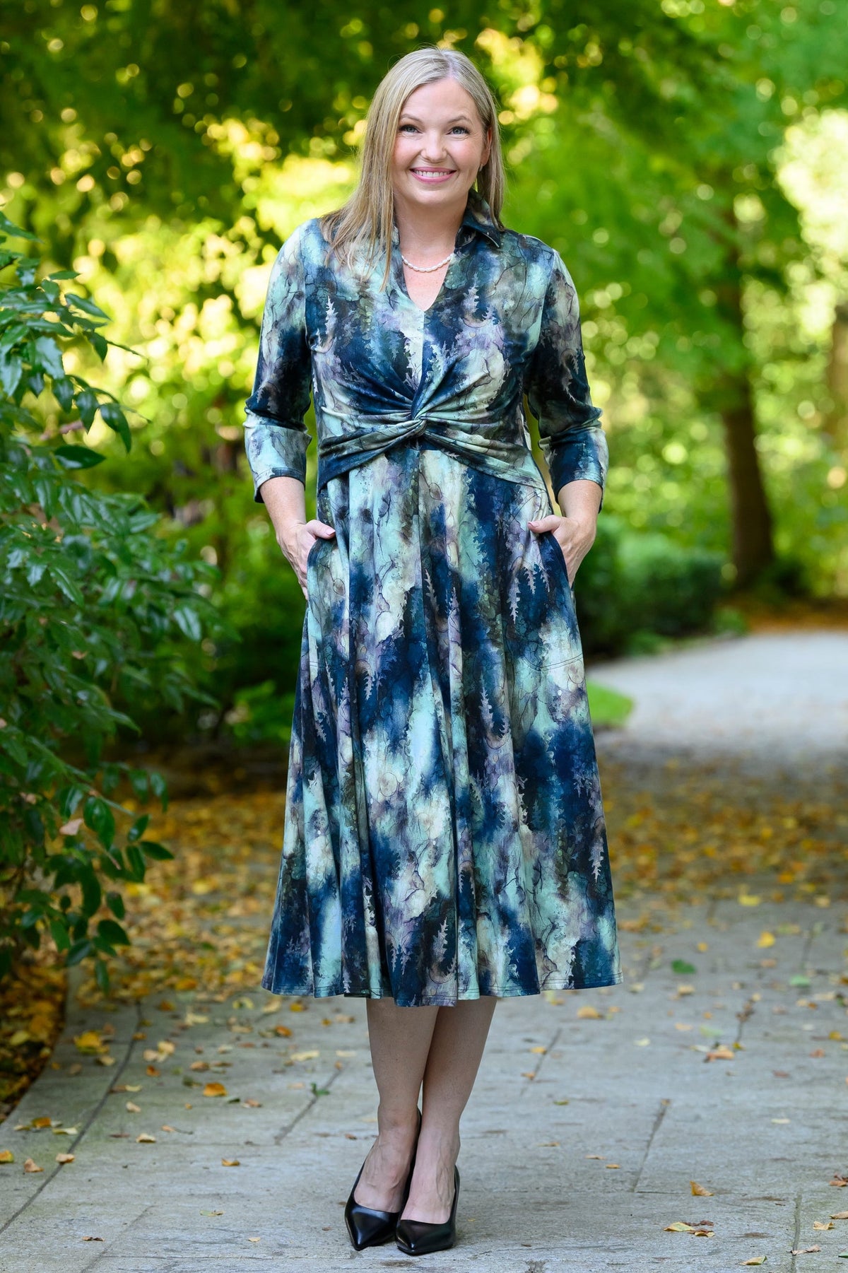 Front view of female model wearing a long-sleeve midi dress with a blue, green, and white tie-dye pattern, standing on a paved path in a park with trees in the background.