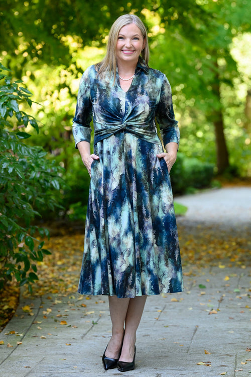Front view of female model wearing a long-sleeve midi dress with a blue, green, and white tie-dye pattern, standing on a paved path in a park with trees in the background.