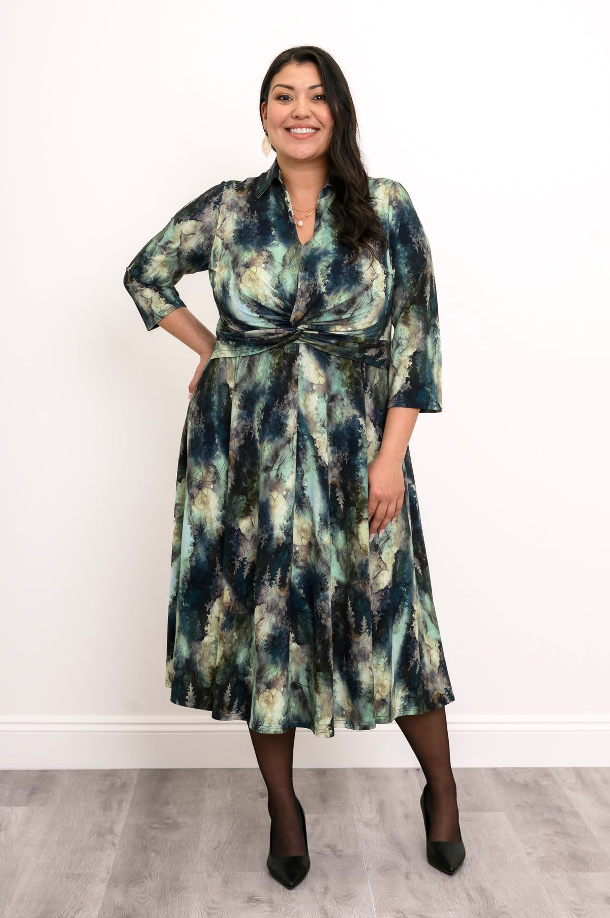 Plus-size model in a green forest print midi dress with a twisted waist, sheer black tights, and black heels.
