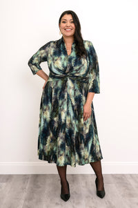 Plus-size model in a green forest print midi dress with a twisted waist, sheer black tights, and black heels.