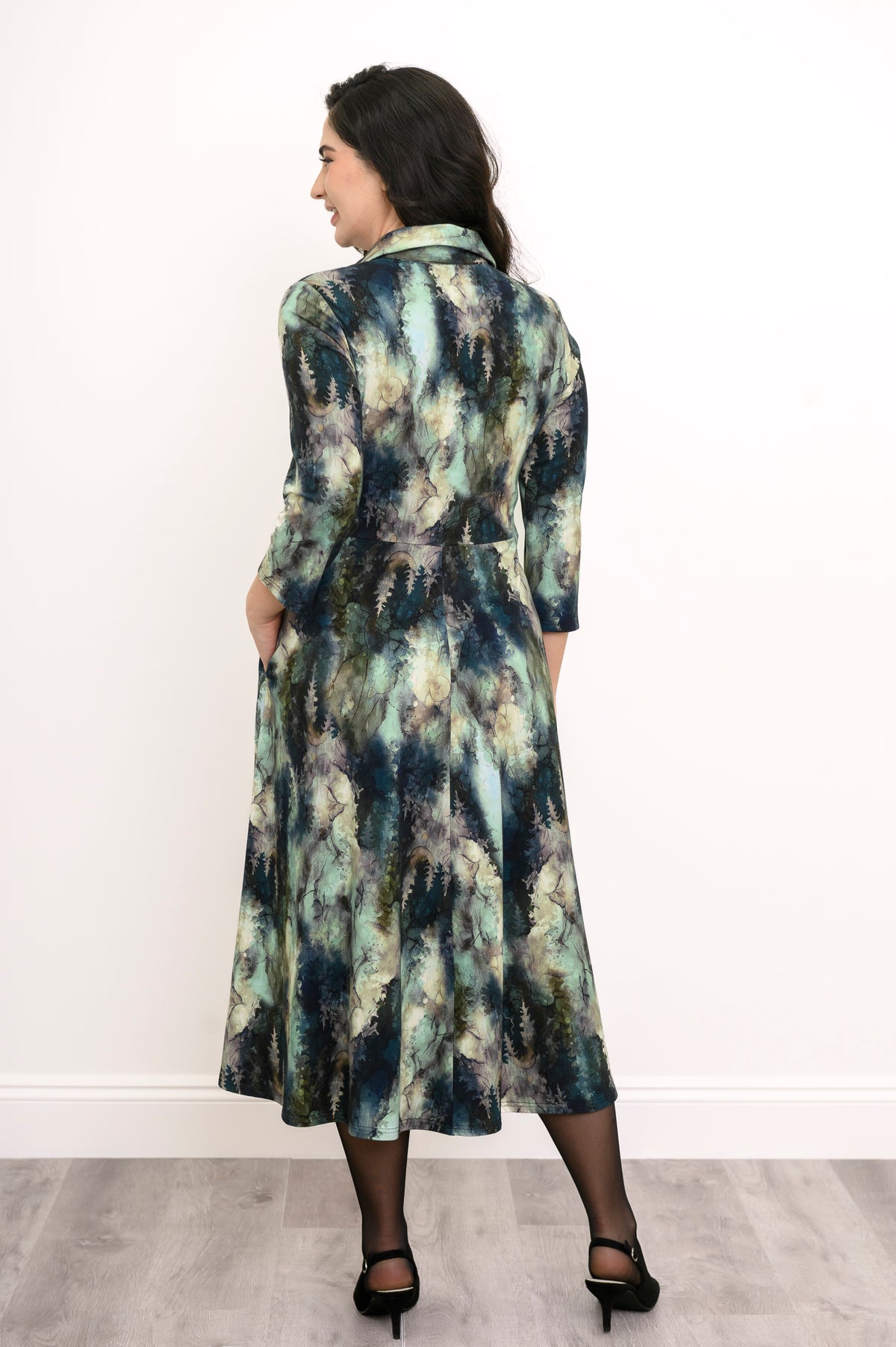 Rear view of a patterned A-line bamboo midi dress with a back seam, styled with sheer tights and black high heels.