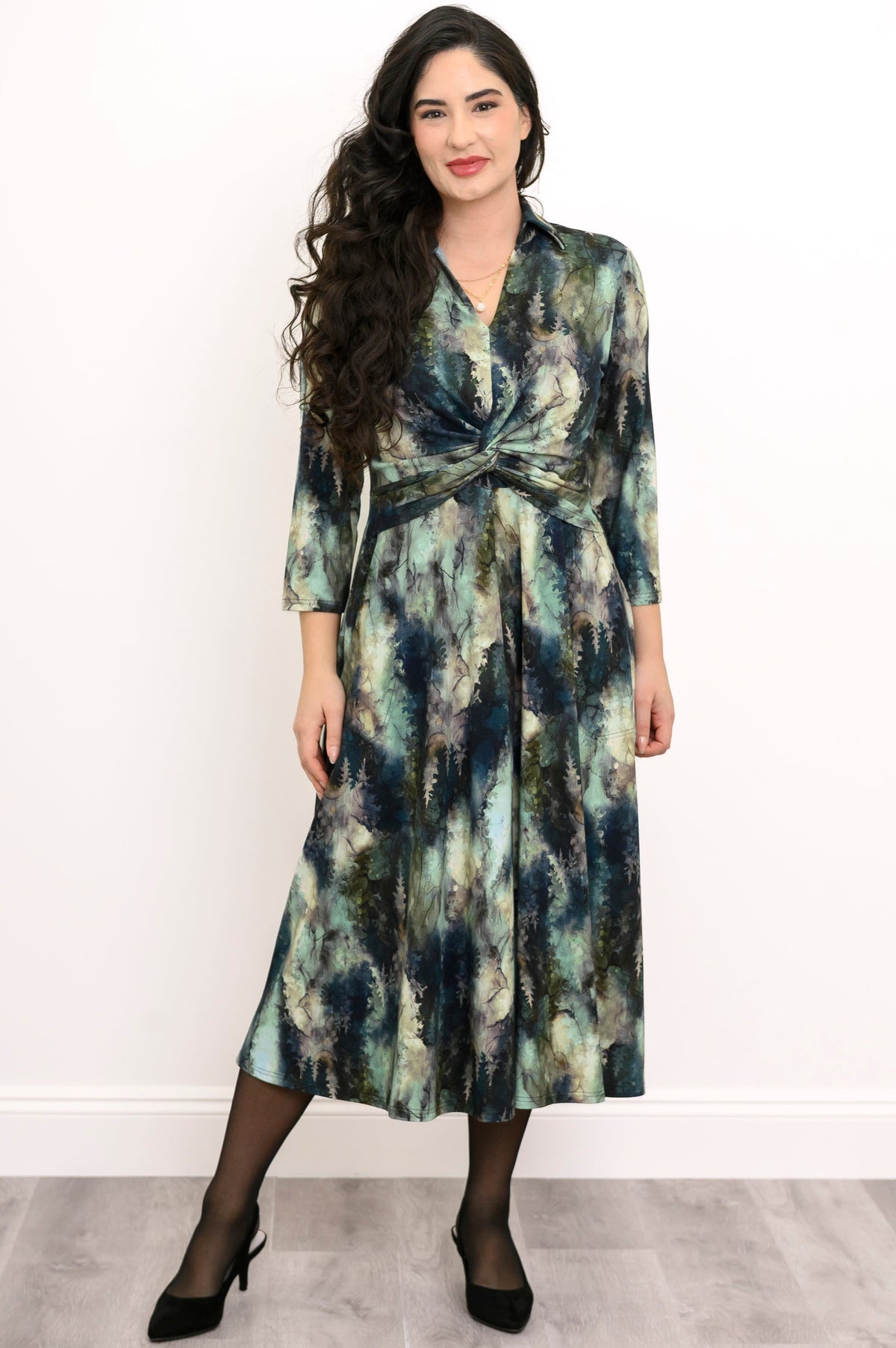 Woman wearing a blue-green watercolor forest print midi dress with a front waist twist, black tights, and heels.