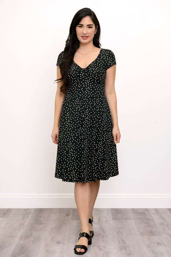 Woman wearing a short sleeve black dress with white polka dots and lack sandals against a white background