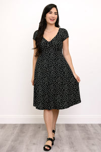 Woman wearing a short sleeve black dress with white polka dots and lack sandals against a white background