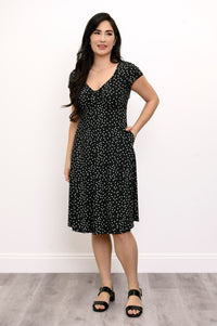 Woman wearing a short sleeve black dress with white polka dots and lack sandals against a white background