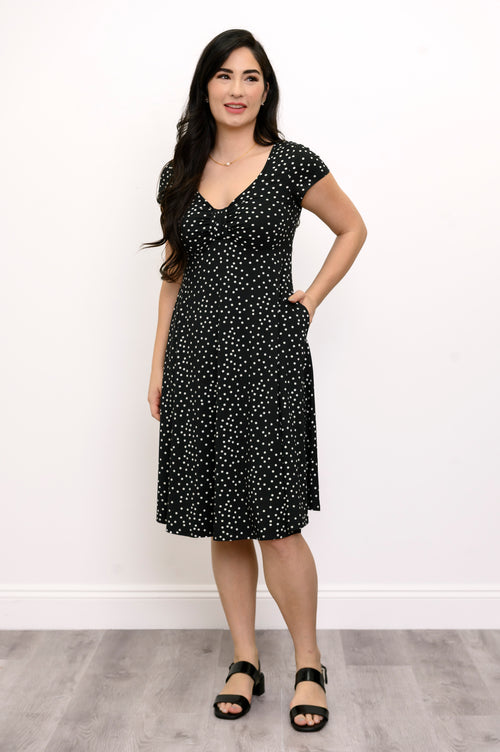 Woman wearing a short sleeve black dress with white polka dots and lack sandals against a white background