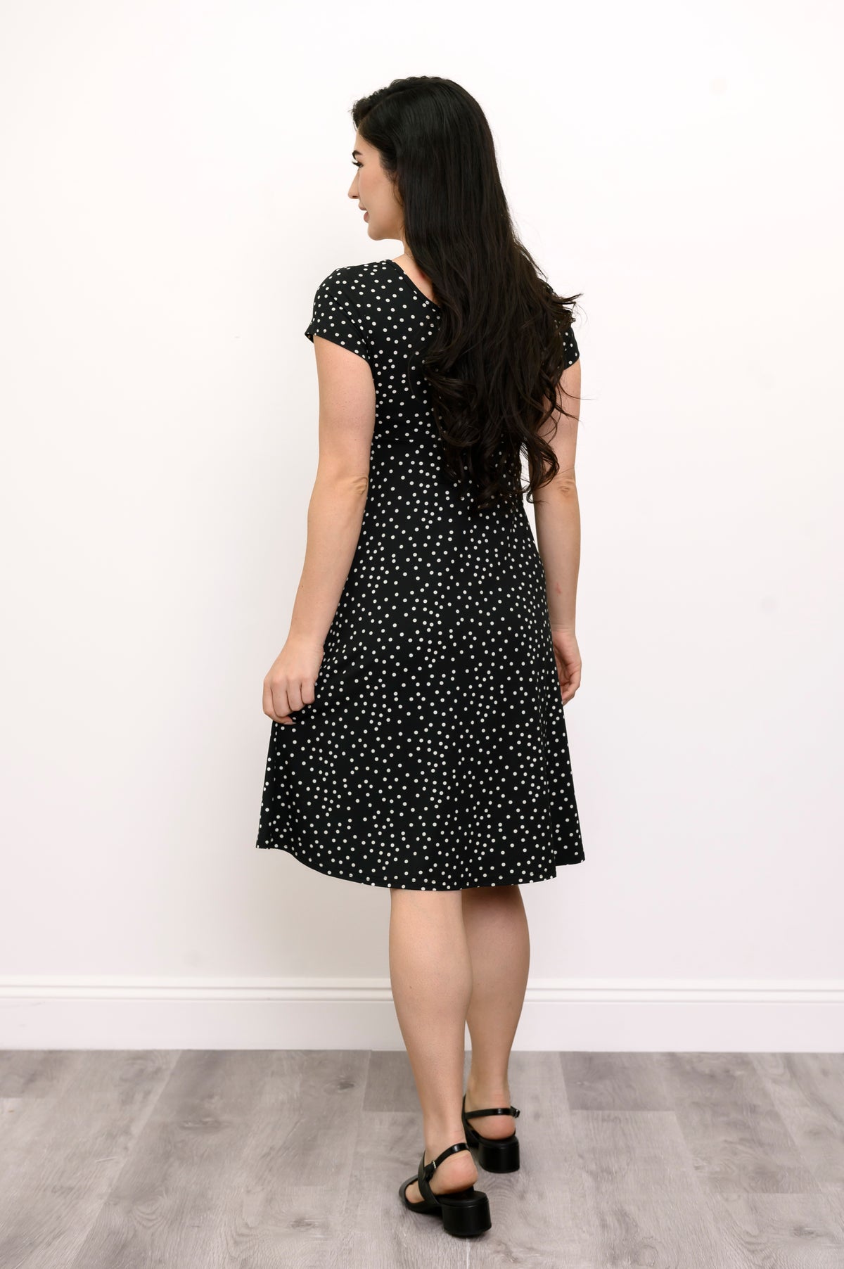 Woman wearing a short sleeve black dress with white polka dots and lack sandals against a white background