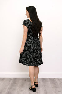 Woman wearing a short sleeve black dress with white polka dots and lack sandals against a white background