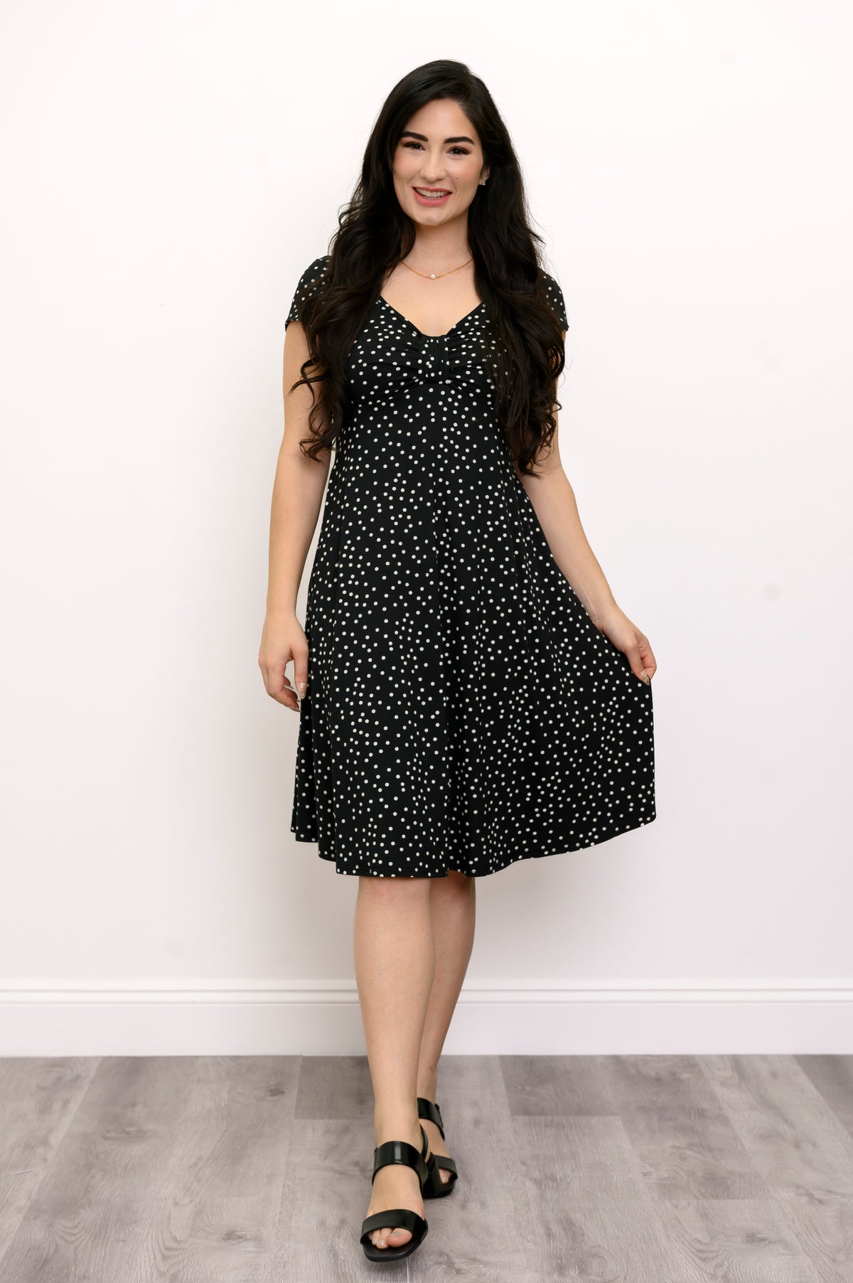 Woman wearing a short sleeve black dress with white polka dots and lack sandals against a white background