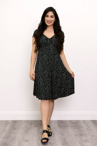 Woman wearing a short sleeve black dress with white polka dots and lack sandals against a white background
