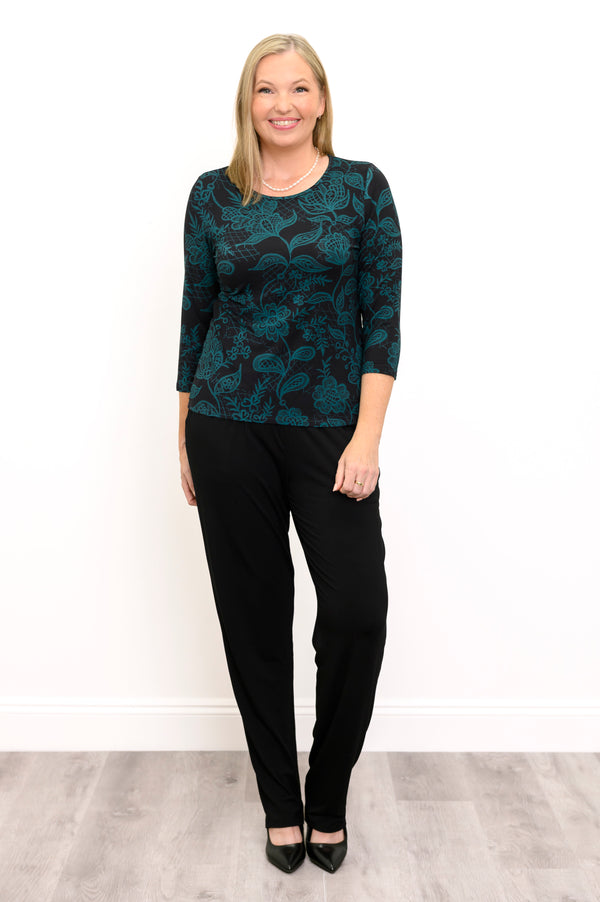 Front view of a female model wearing a 3/4 sleeve top in black with teal floral and leaf print, with black pants and heels against a white backdrop on grey floor