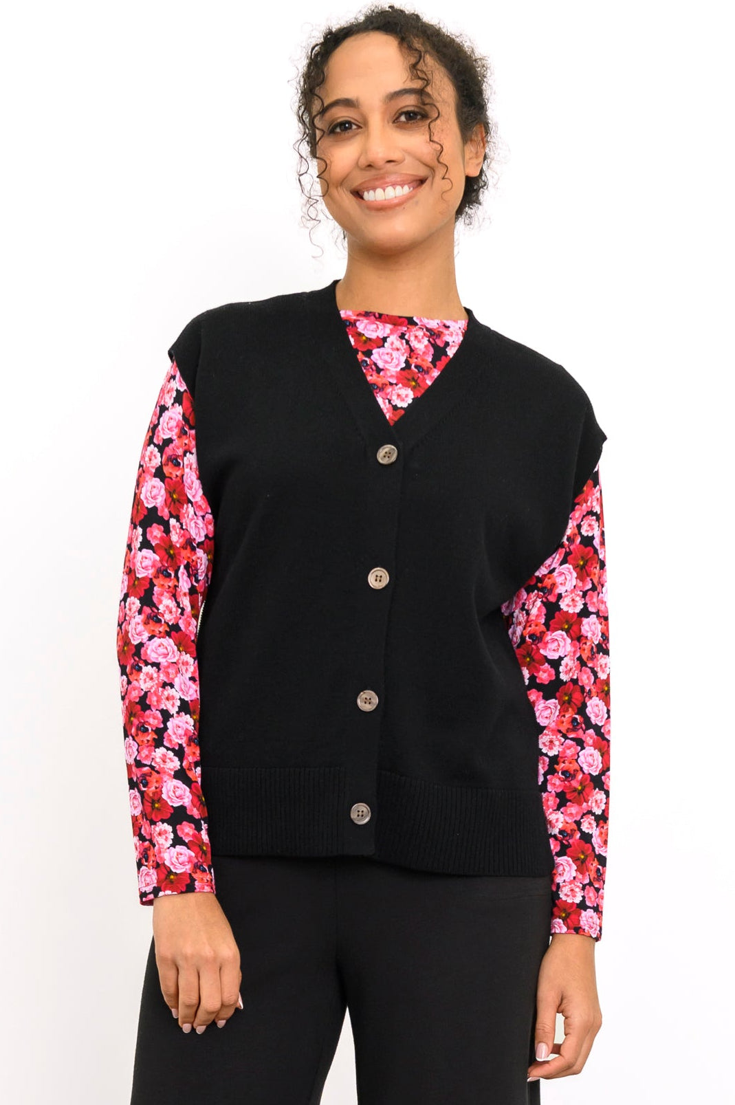 Smiling woman wearing an onyx button-up knit vest layered over a bright pink floral long-sleeve top, front view.