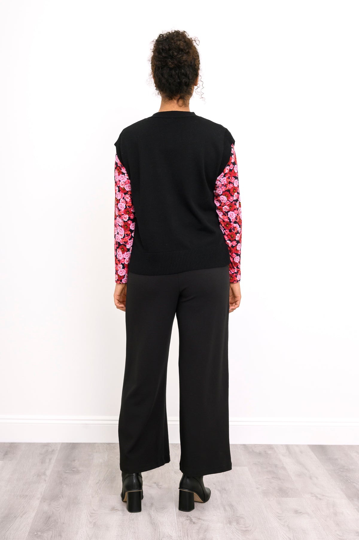 Back view of a women's V-neck black knit vest, styled with a pink patterned shirt and black trousers.