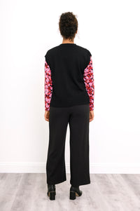 Back view of a women's V-neck black knit vest, styled with a pink patterned shirt and black trousers.