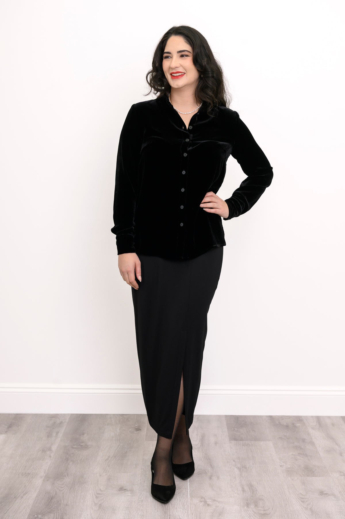Wide studio shot of an elegant women's black velvet long-sleeve blouse paired with a long black skirt and heels