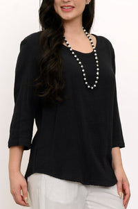 Woman wearing a black top and white pants with a beaded necklace against a white wall, close-up front view