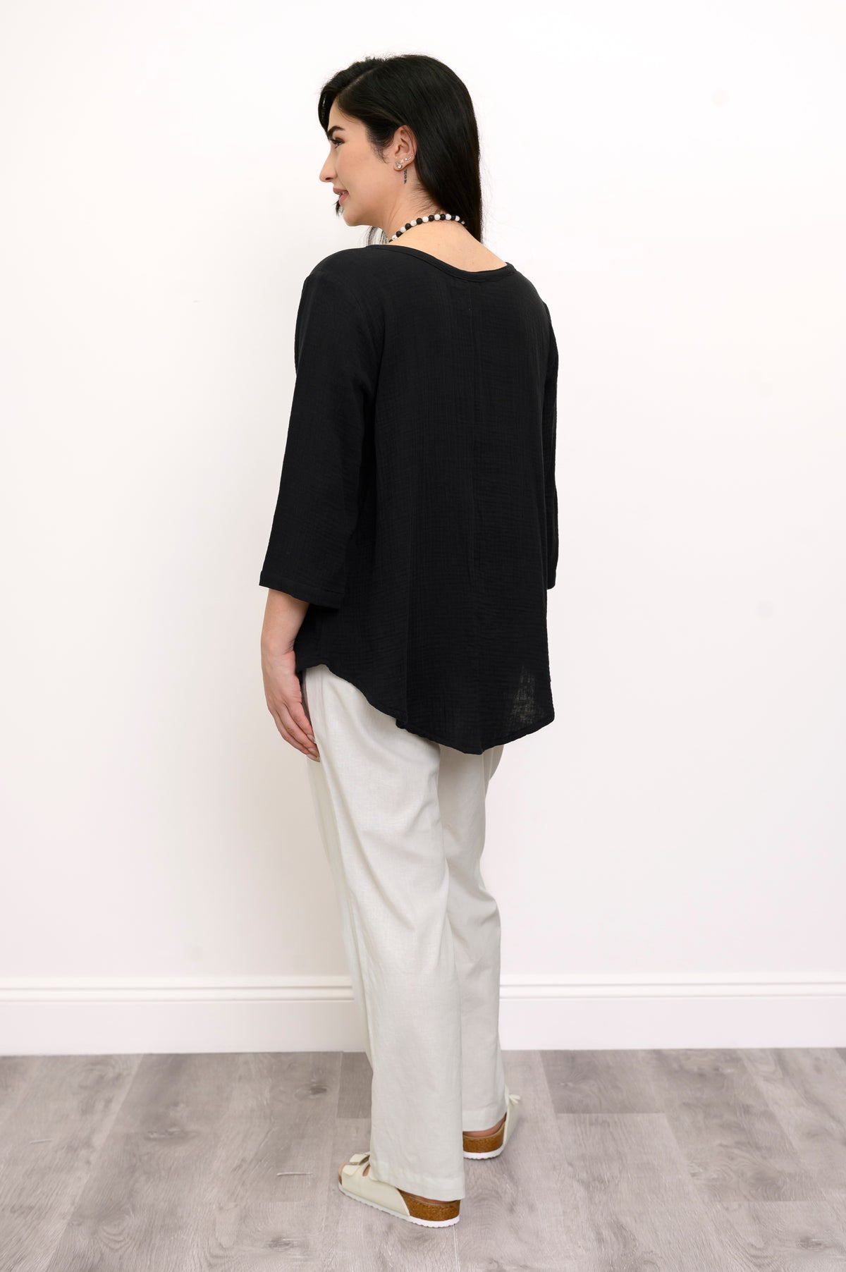 Woman wearing a black top and white pants with a beaded necklace against a white wall, back view