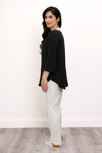Woman wearing a black top and white pants with a beaded necklace against a white wall, side view