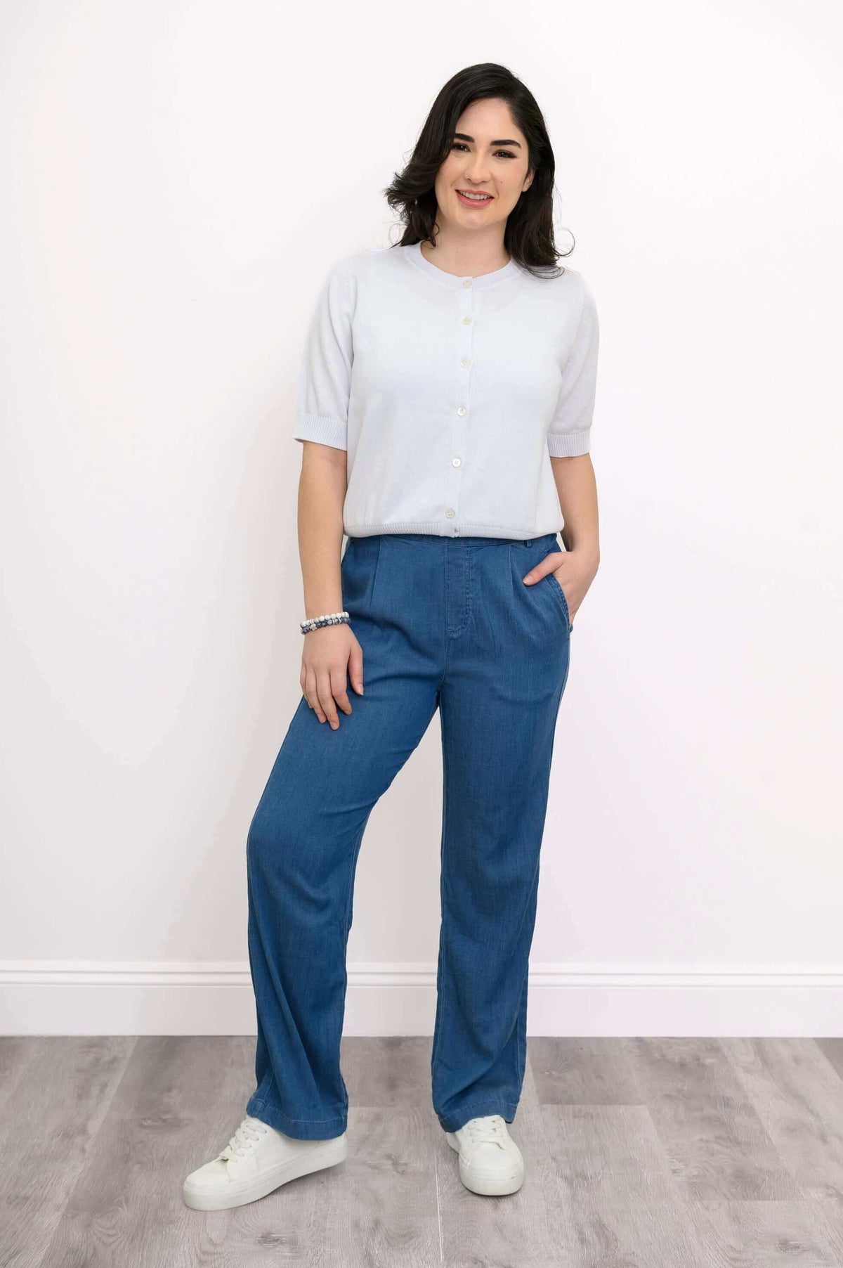Model wearing a button-up white cardigan with blue pants, hand in pocket