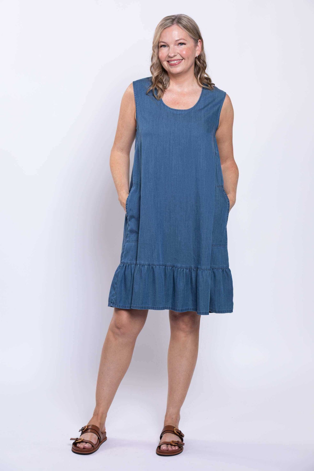 Model smiling in sleeveless blue dress, hands in pockets, with ruffle hem
