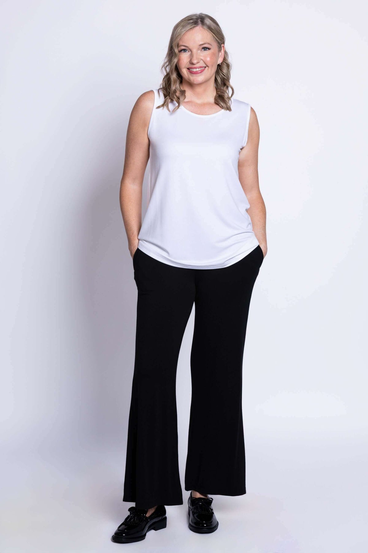 Model in white tank top with hands in pockets, wearing black pants
