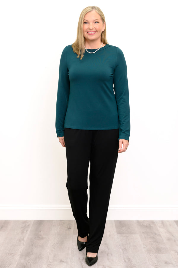 Front view of a female model in walking pose wearing a teal long sleeve top with black pants and shoes, against a white wall and gray floor