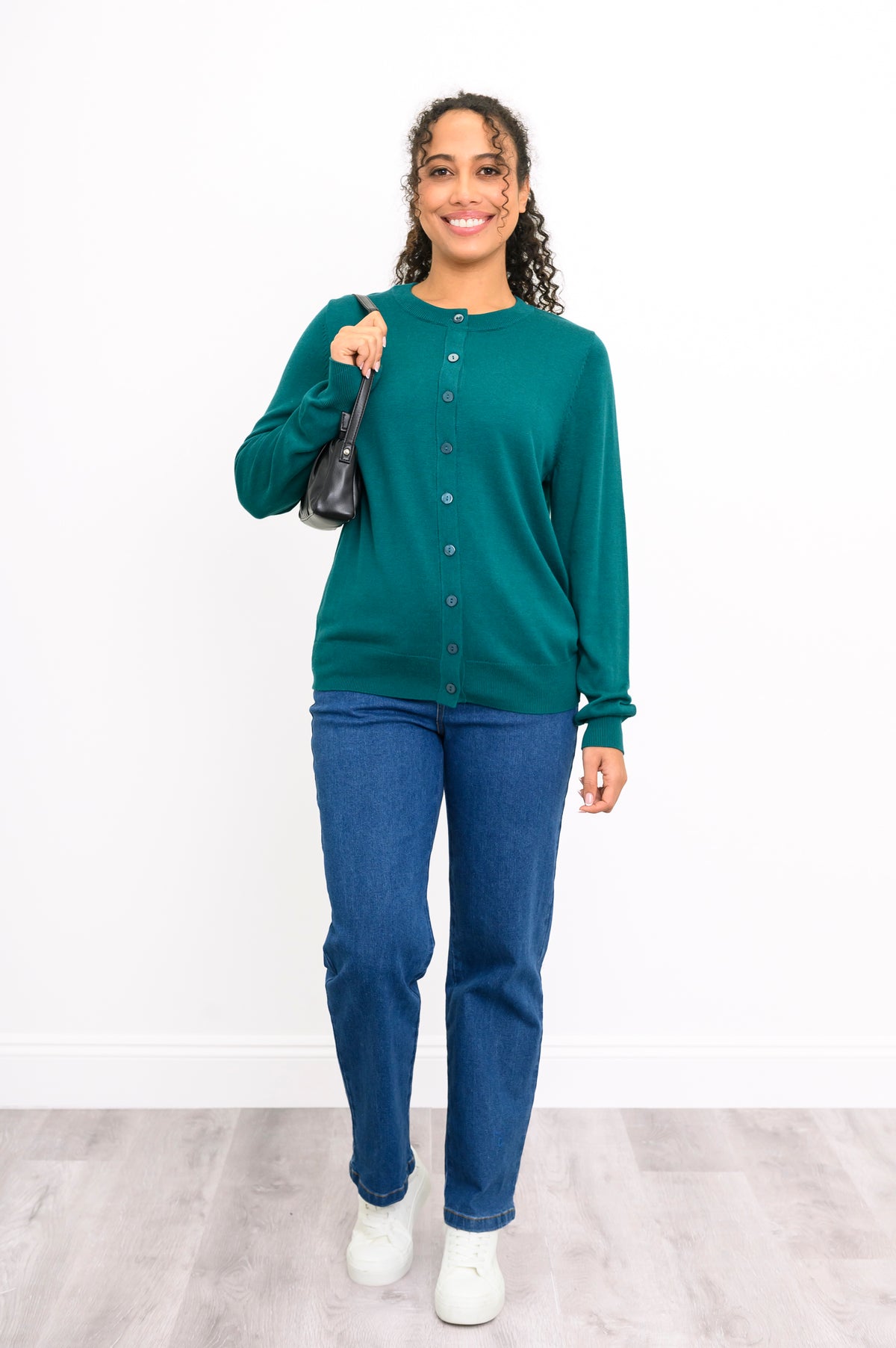 Full length shot of a woman in a teal button down knit top with a black crossbody purse and jeans.