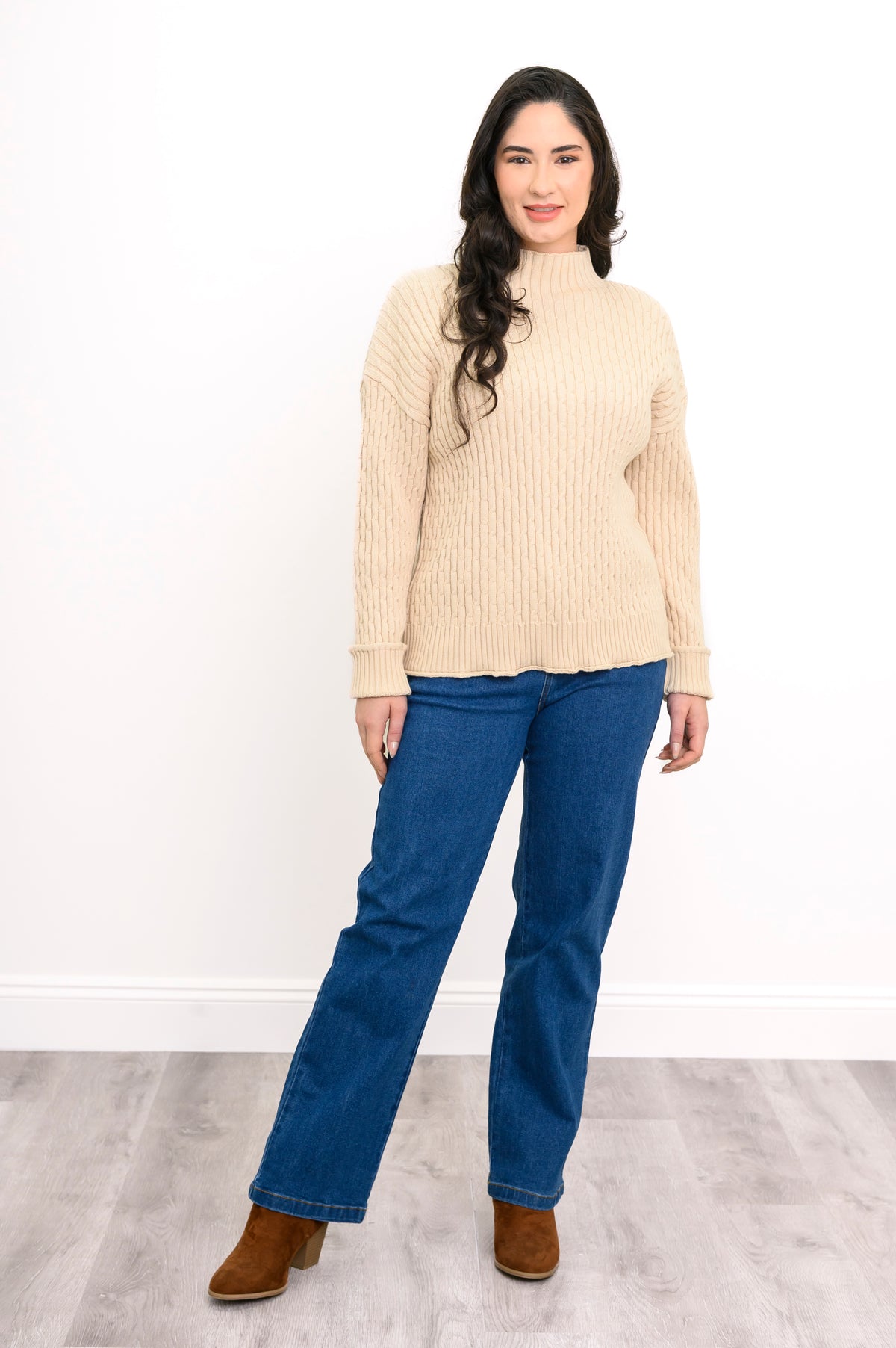 Model posing in a cream mock neck knit sweater, blue denim jeans, and brown leather boots, front-facing view.