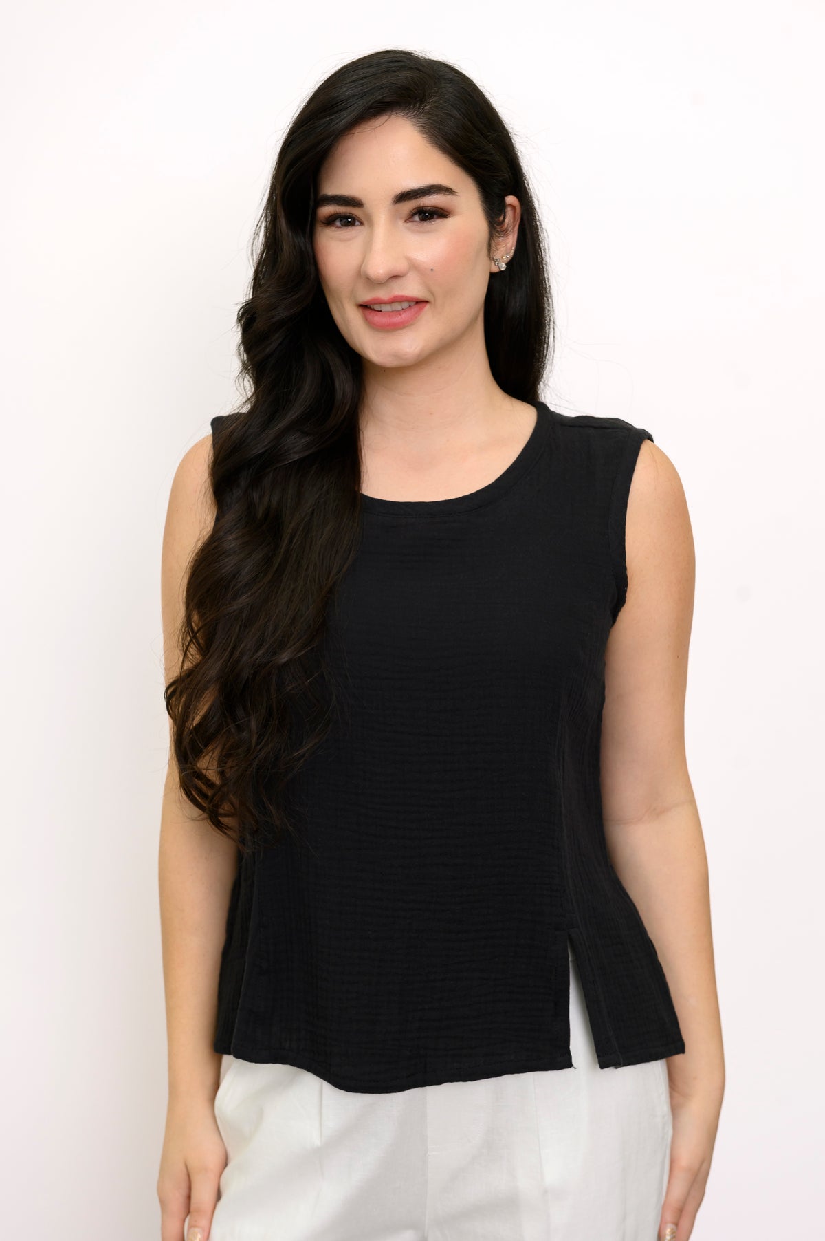 Woman wearing a black sleeveless top and white pants against a white background, front view